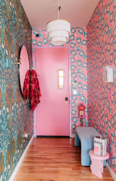 This is a stylish entryway with colorful wallpaper. The left wall has blue and brown patterned wallpaper, while the right wall has pink wallpaper with blue shapes. A round mirror hangs on the left wall. A pink door is straight ahead. A three-tiered white light fixture hangs from the ceiling. A small blue bench and pink stool are on the right.